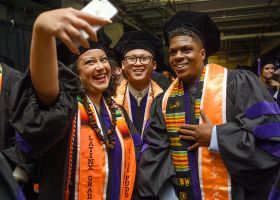 Three students dressed in graduation regalia take a selfie 