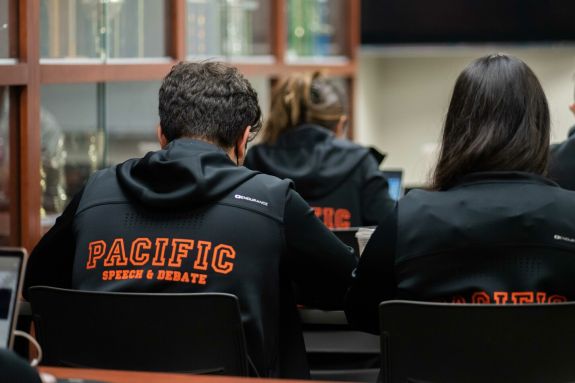 Students wearing their Pacific Speech and Debate sweatshirts are sitting at tables in their team room. Trophies are visible in the cases next to them.